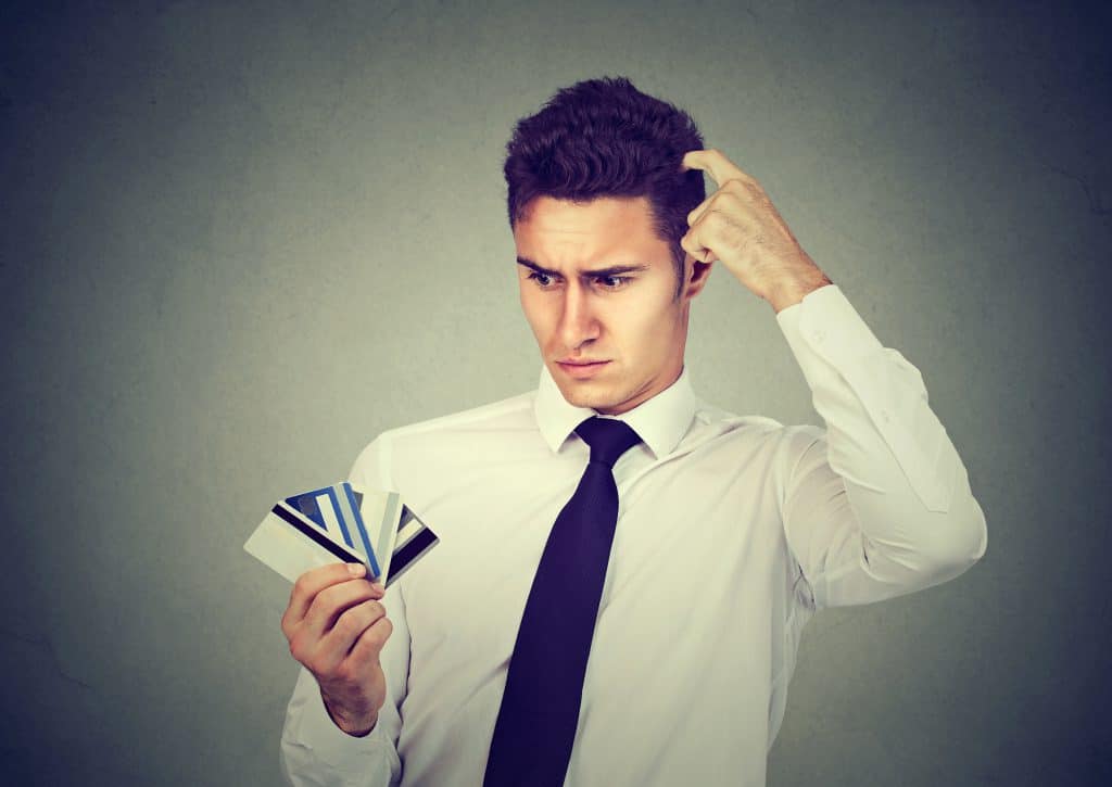 Confused man looking at many credit cards uncertain which one to choose isolated on gray background